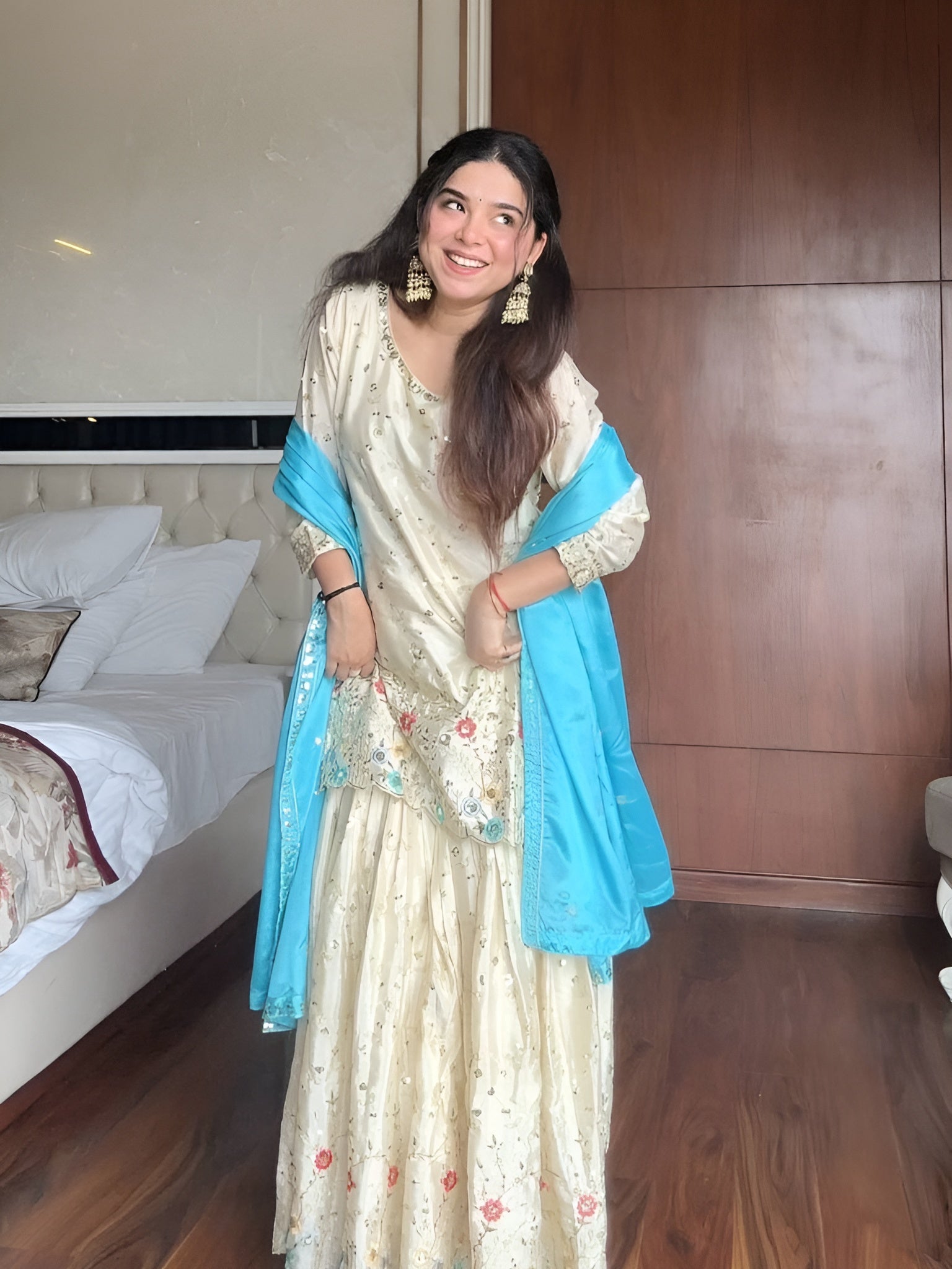 Woman in a cream & blue embroidered chinon sharara suit standing in a bedroom with wooden walls and a bed.