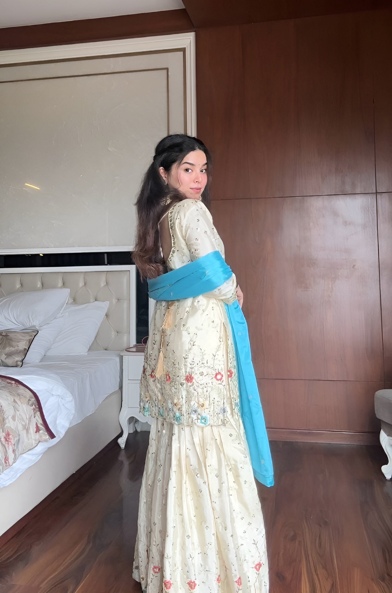 Woman in a cream & blue embroidered chinon sharara suit standing in a bedroom with wooden walls and a bed.