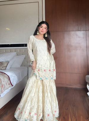 Woman in a cream & blue embroidered chinon sharara suit standing in a bedroom with wooden walls and a bed.