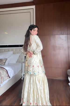 Woman in a cream & blue embroidered chinon sharara suit standing in a bedroom with wooden walls and a bed.