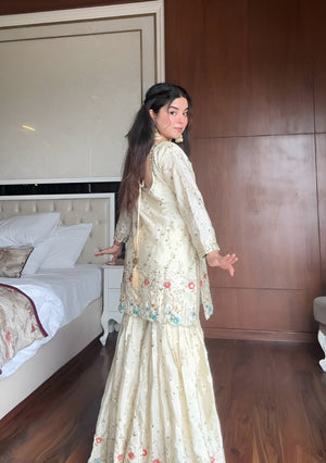 Woman in a cream & blue embroidered chinon sharara suit standing in a bedroom with wooden walls and a bed.