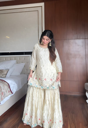 Woman in a cream & blue embroidered chinon sharara suit standing in a bedroom with wooden walls and a bed.