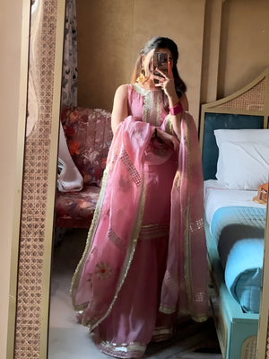 Person wearing a pink traditional Diya Organza Sharara Set with Sequin_Mirror Work outfit in a room with a bed and sofa.Lahariya.com