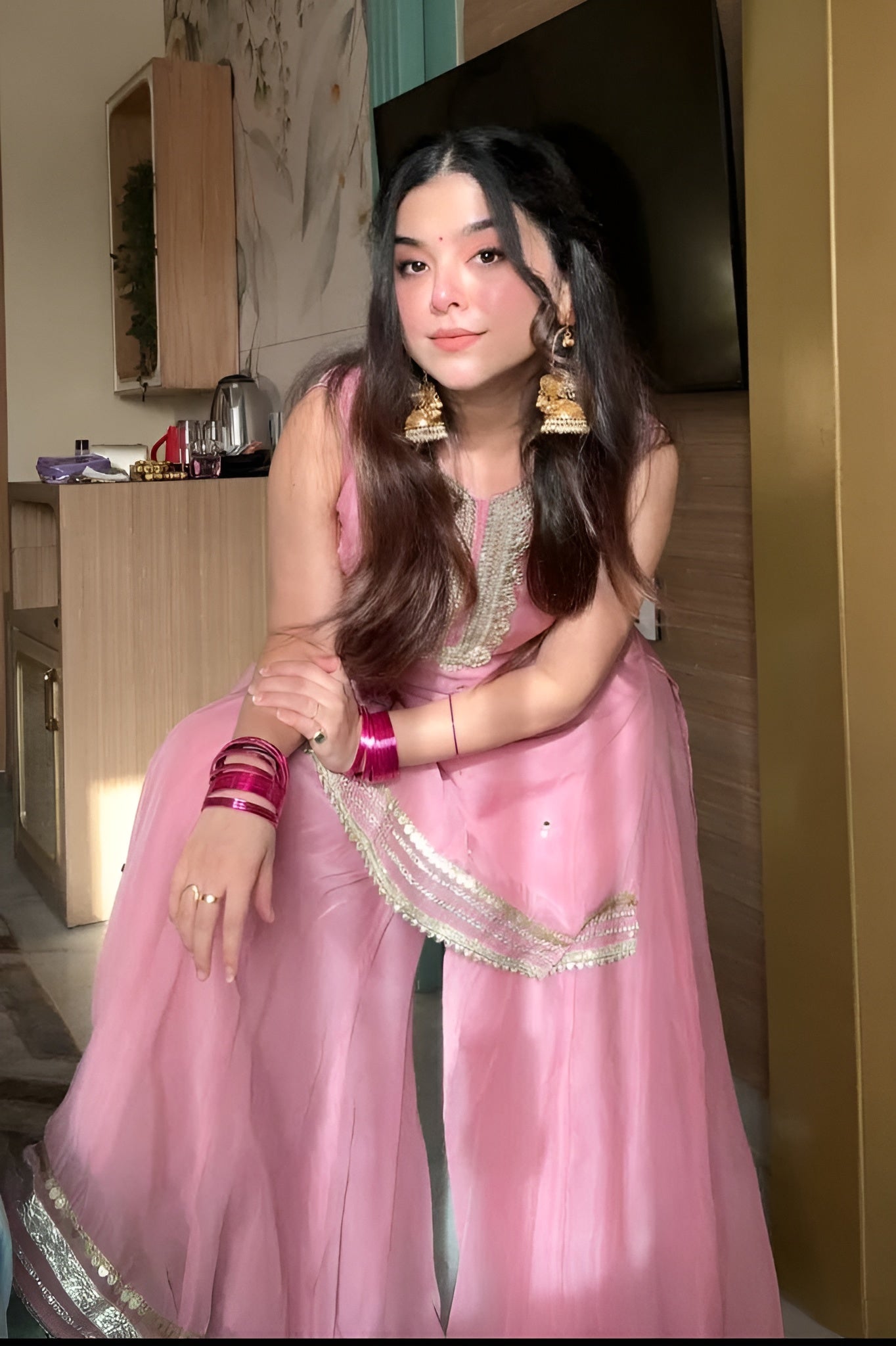 Person wearing a pink traditional Diya Organza Sharara Set with Sequin_Mirror Work outfit in a room with a bed and sofa.Lahariya.com
