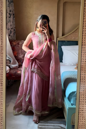 Person wearing a pink traditional Diya Organza Sharara Set with Sequin_Mirror Work outfit in a room with a bed and sofa.Lahariya.com