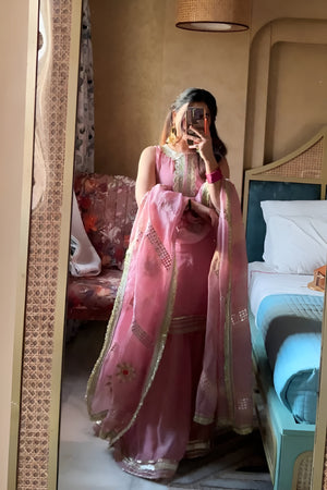 Person wearing a pink traditional Diya Organza Sharara Set with Sequin_Mirror Work outfit in a room with a bed and sofa.Lahariya.com
