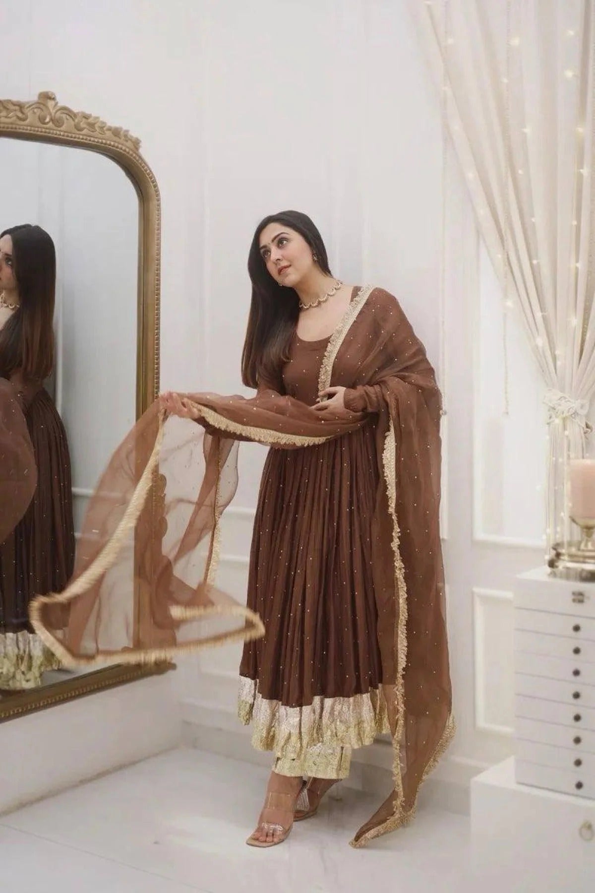 Woman in a brown traditional outfit with a sheer dupatta, standing in front of a mirror.