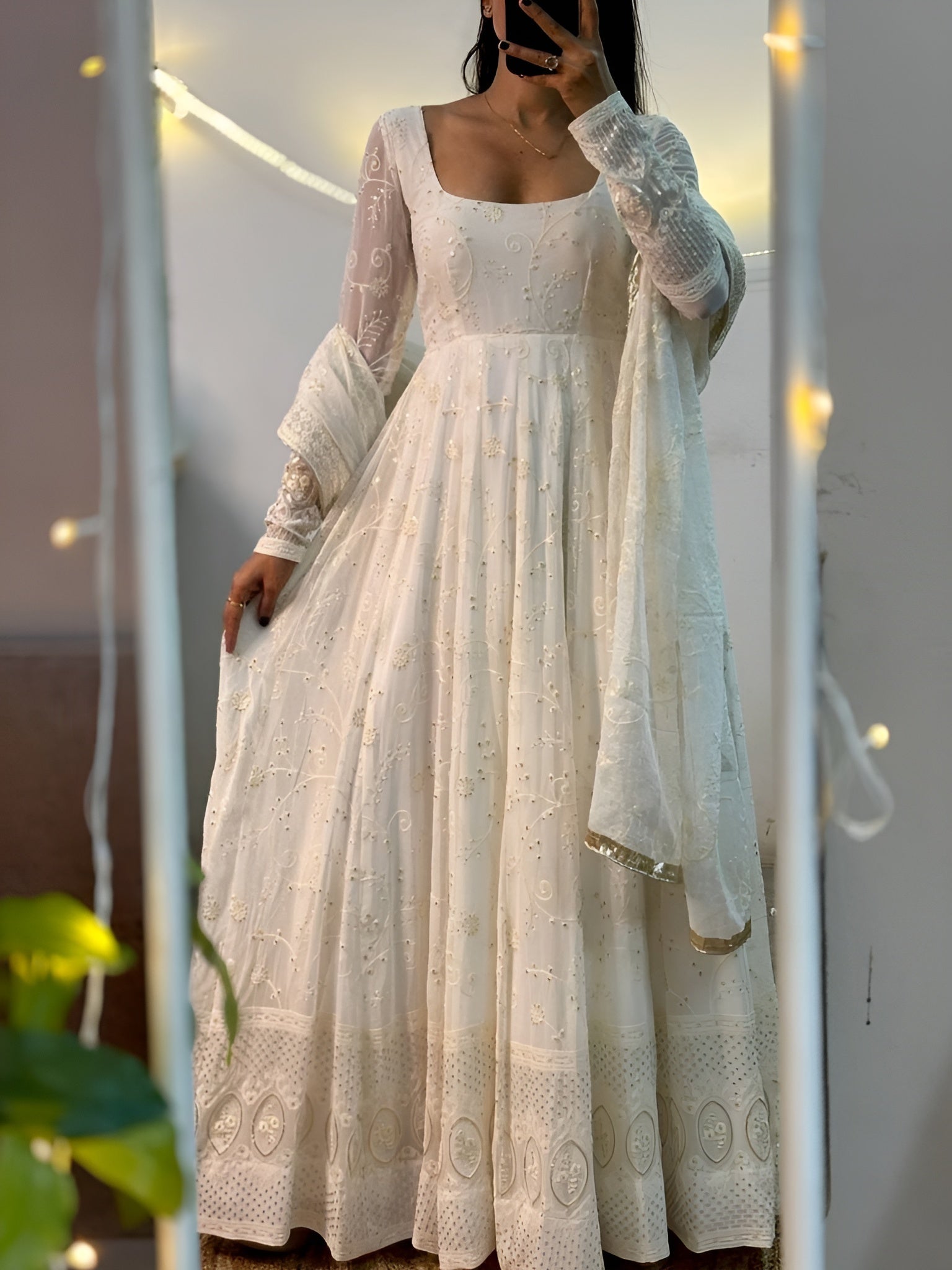 Woman wearing a white embroidered dress with a sheer dupatta, standing indoors with string lights in the background.