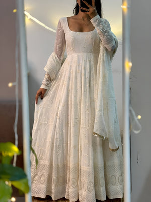 Woman wearing a white embroidered dress with a sheer dupatta, standing indoors with string lights in the background.