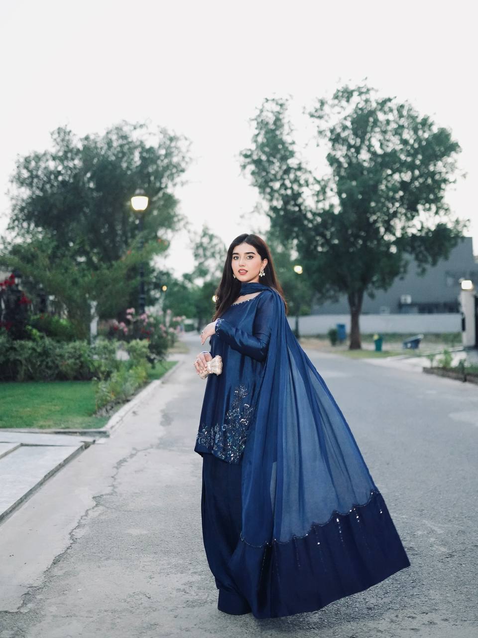 Woman in a blue traditional outfit standing on a street with trees in the background