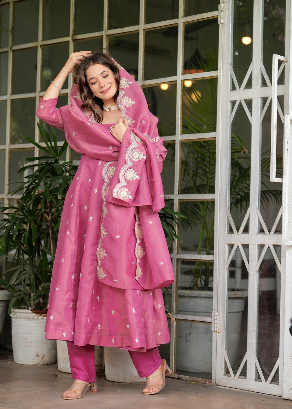 Woman in a pink traditional outfit Riya Pink Georgette Kurti Pant Set with Embroidered Dupatta standing in front of a glass door with plants.