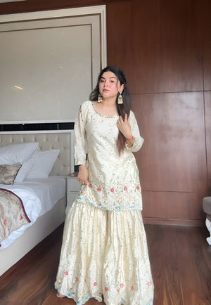 Woman in a cream & blue embroidered chinon sharara suit standing in a bedroom with wooden walls and a bed.