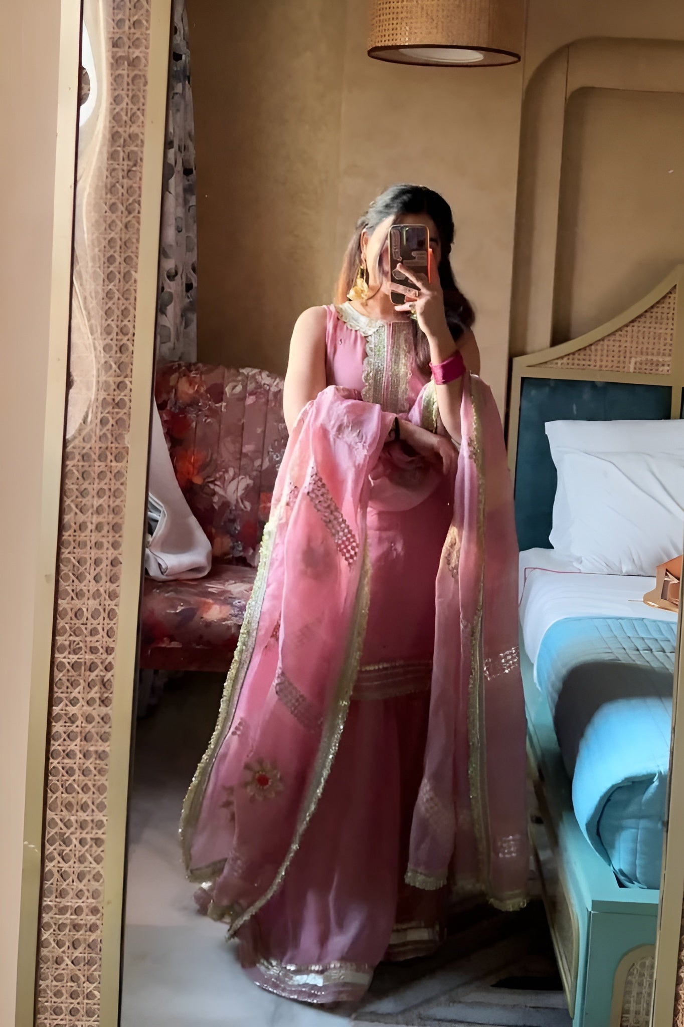 Person wearing a pink traditional Diya Organza Sharara Set with Sequin_Mirror Work outfit in a room with a bed and sofa.Lahariya.com