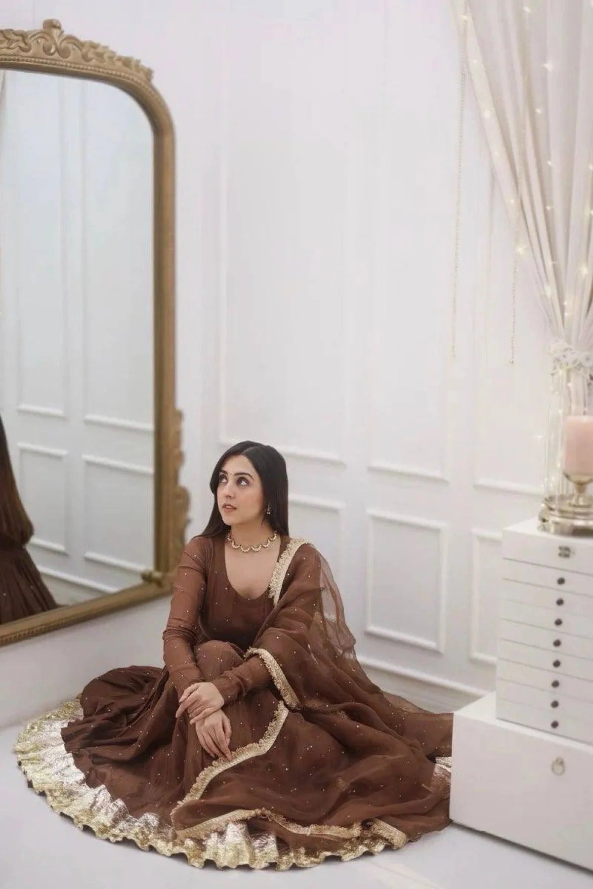Woman in a brown dress sitting in front of a mirror in a room with white walls and decor.