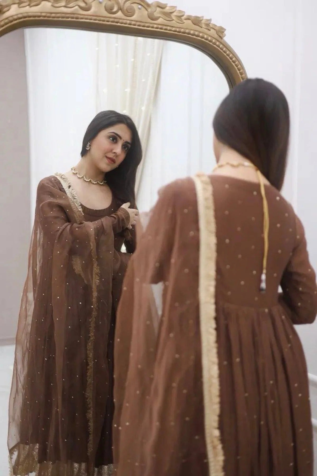 Woman in a brown dress with gold details looking at herself in a mirror.