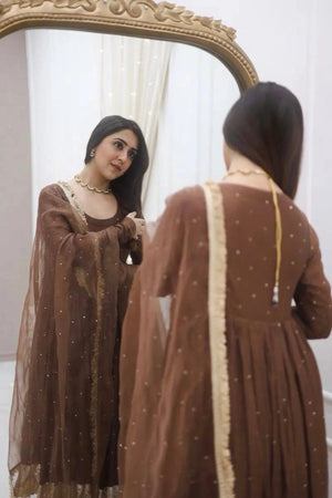 Woman in a brown dress with gold details looking at herself in a mirror.