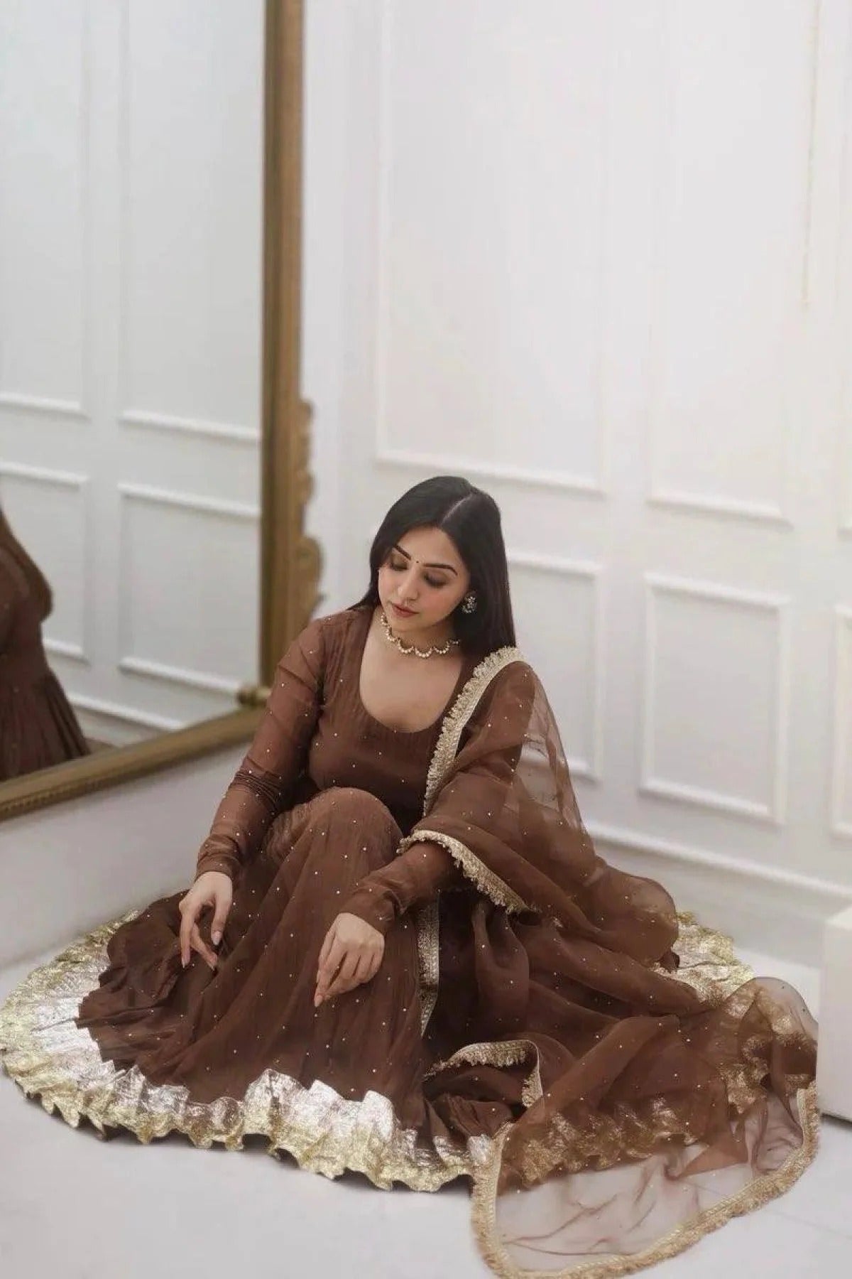 Woman in a brown traditional outfit sitting on a white floor with a mirror in the background.