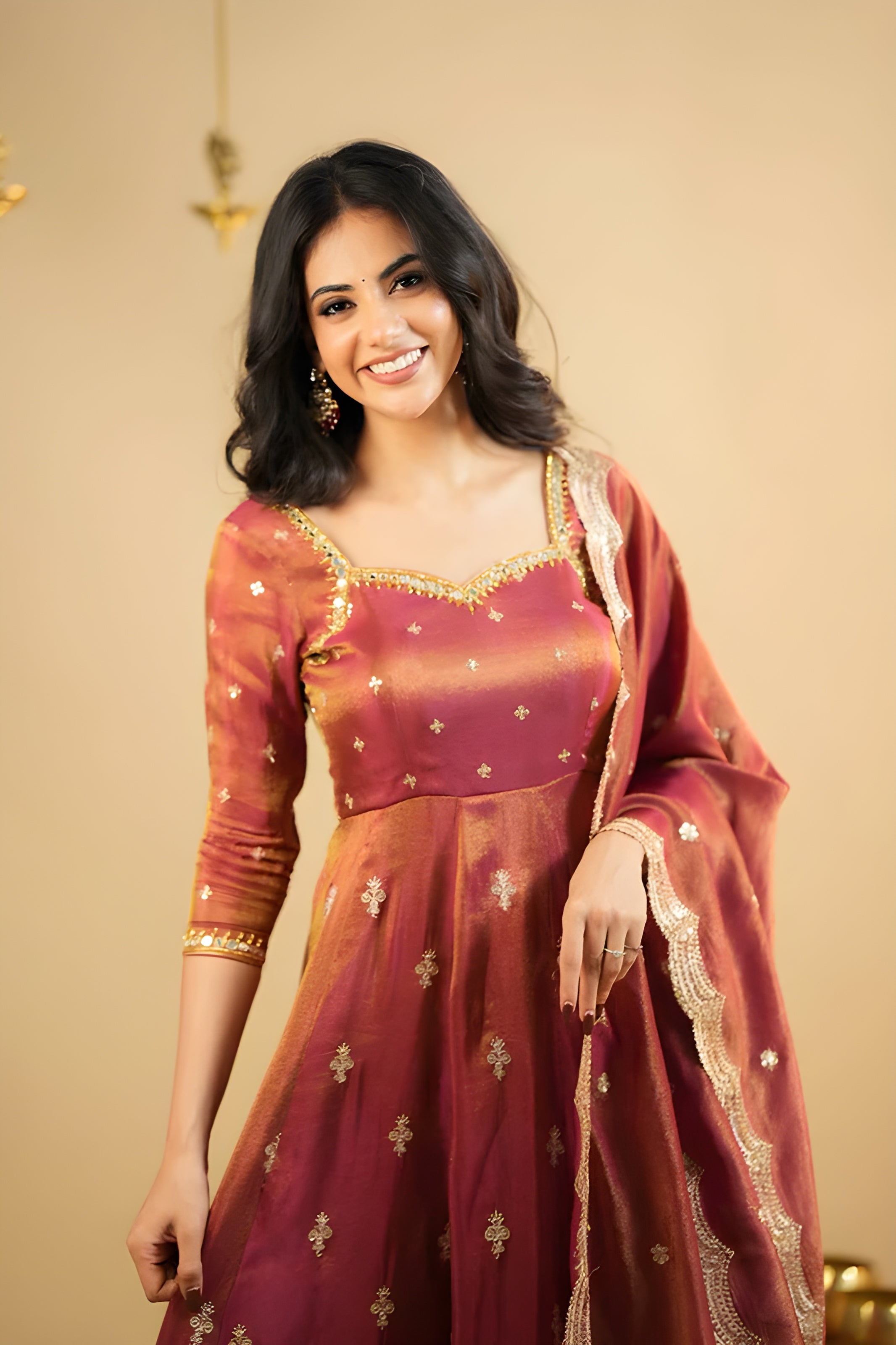 Woman wearing a maroon and orange traditional outfit with gold embroidery against a beige background
