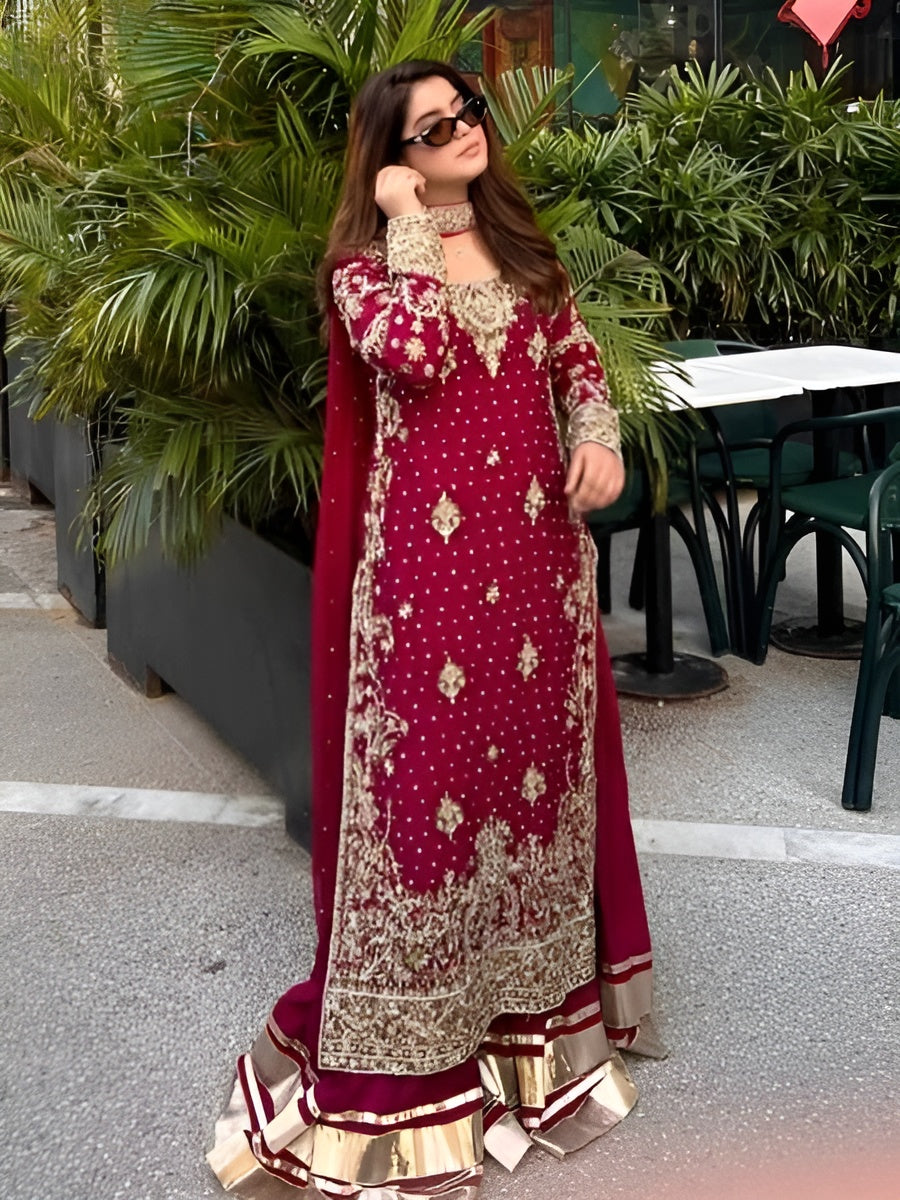 Woman in a red and gold traditional outfit standing outdoors with greenery in the background