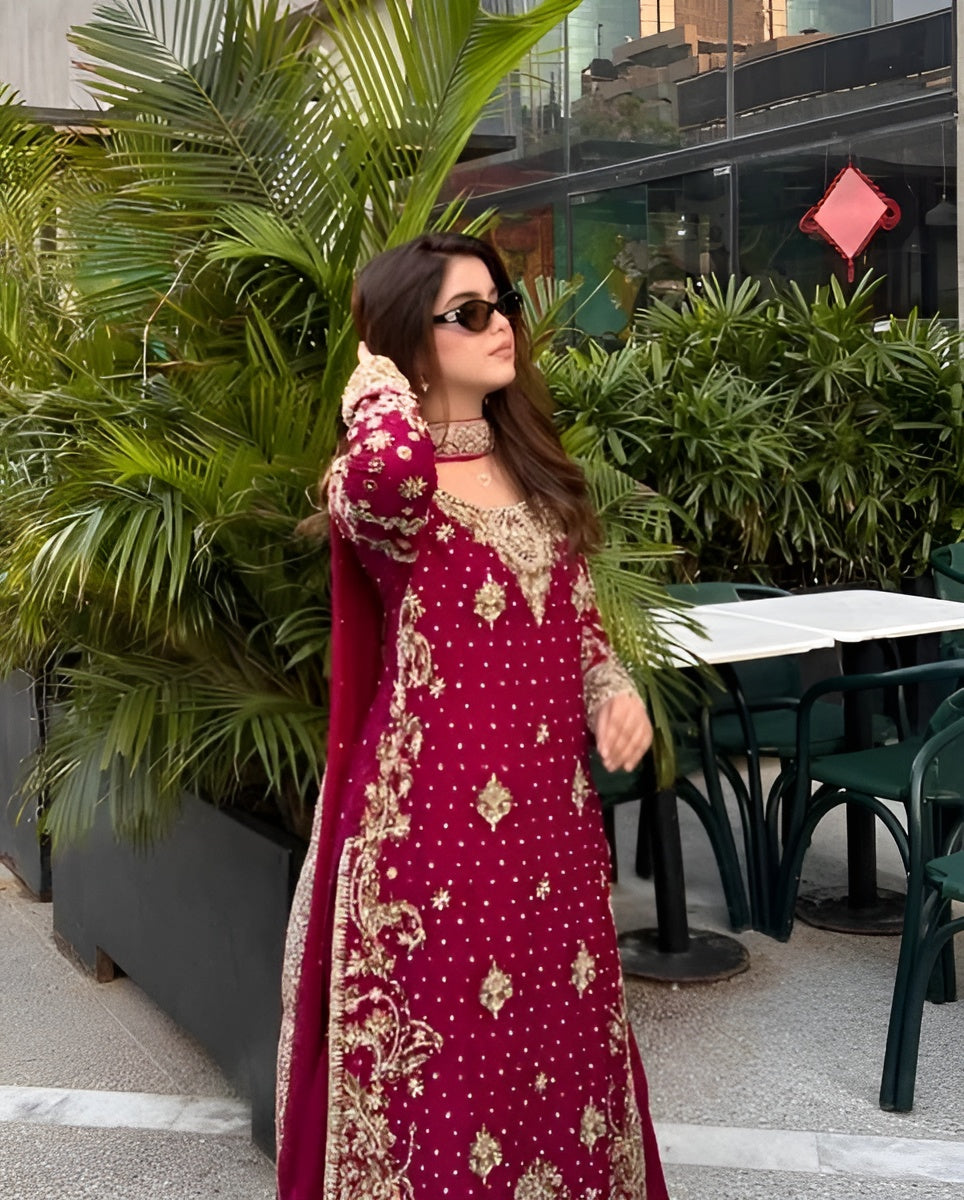 Woman in a red and gold embroidered dress standing outdoors with greenery and tables in the background.