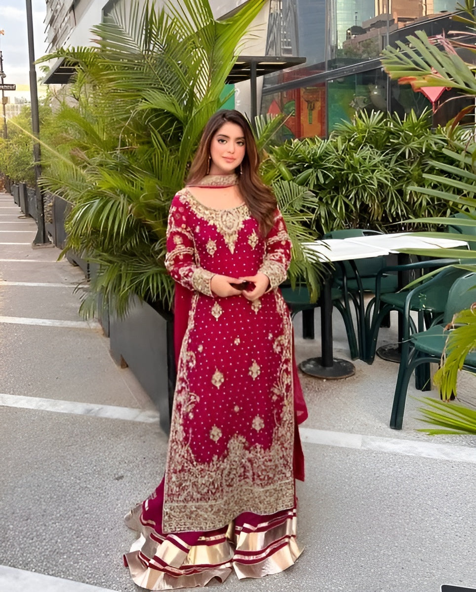 Woman in a red and gold traditional outfit standing outdoors with greenery in the background