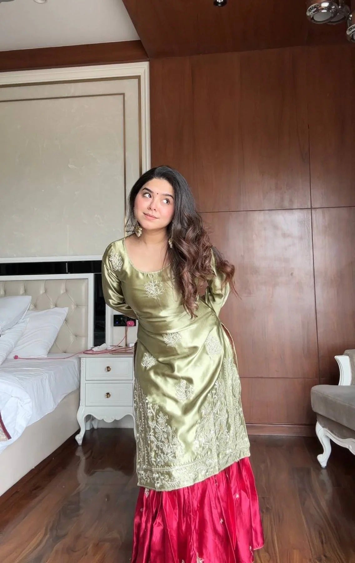 Woman in a green and red traditional outfit standing in a bedroom.