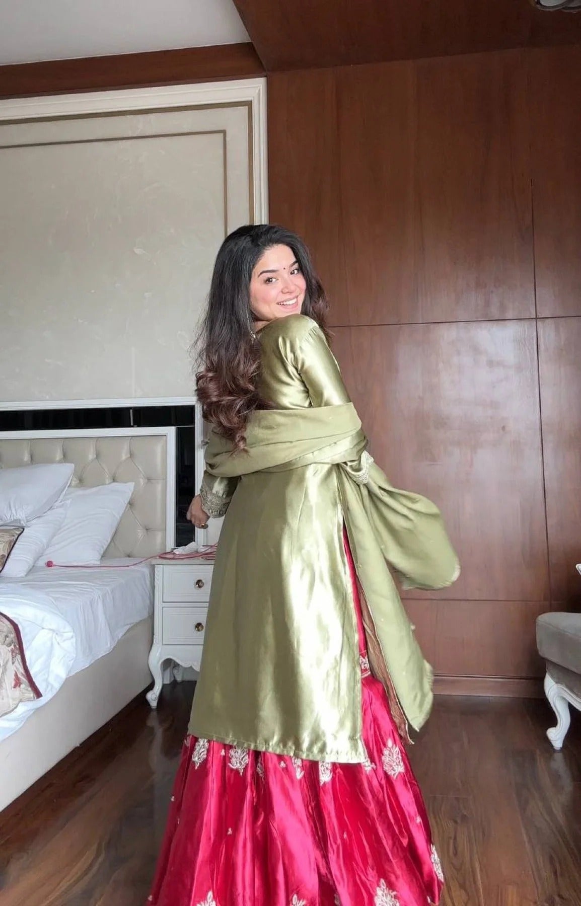 Woman in a green and red traditional outfit standing in a bedroom.