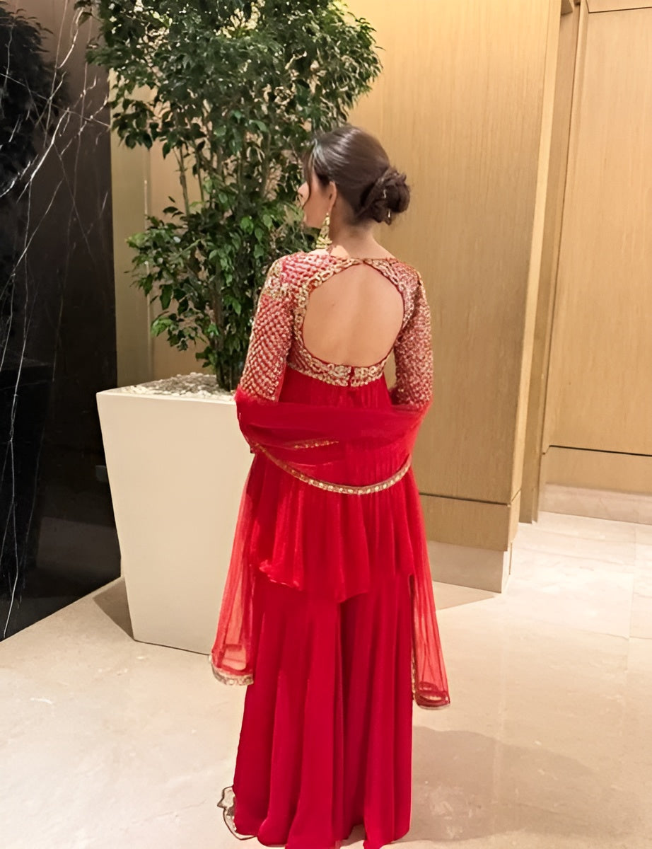 Woman in a red dress with a textured top and sheer overlay standing indoors.
