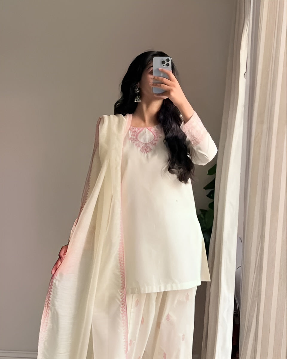 Woman wearing a light-colored traditional outfit with pink embroidery, standing in front of a mirror.