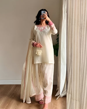 Person wearing a traditional outfit with embroidery, standing in a room with curtains.