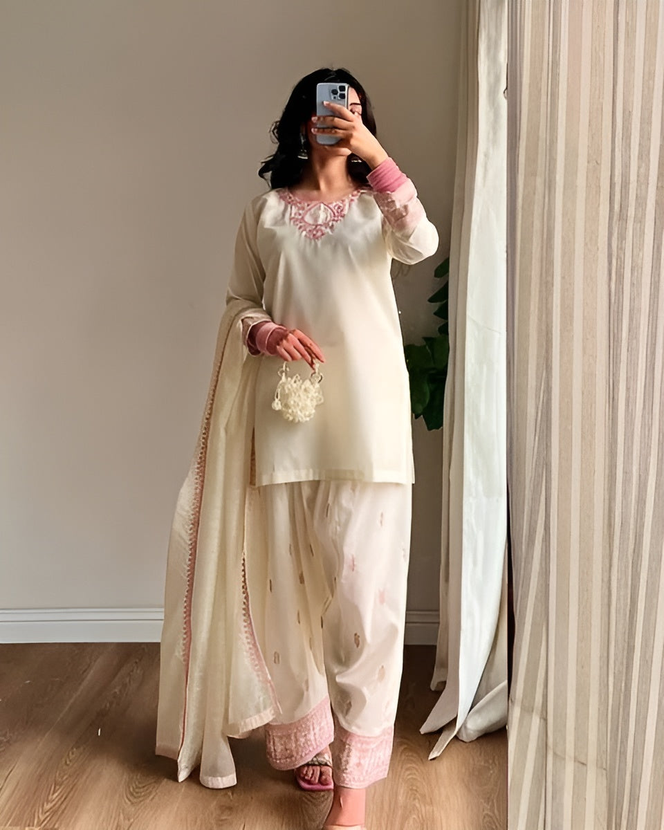Person wearing a traditional outfit with embroidery, standing in a room with curtains.