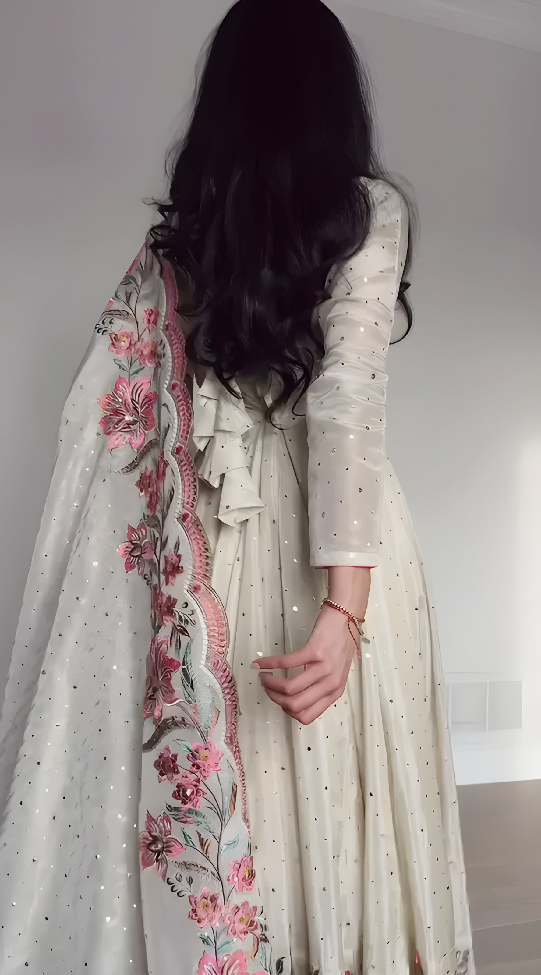 Person wearing a long, light-colored dress with floral embroidery in a minimal indoor setting.
