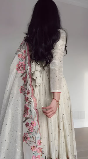 Person wearing a long, light-colored dress with floral embroidery in a minimal indoor setting.