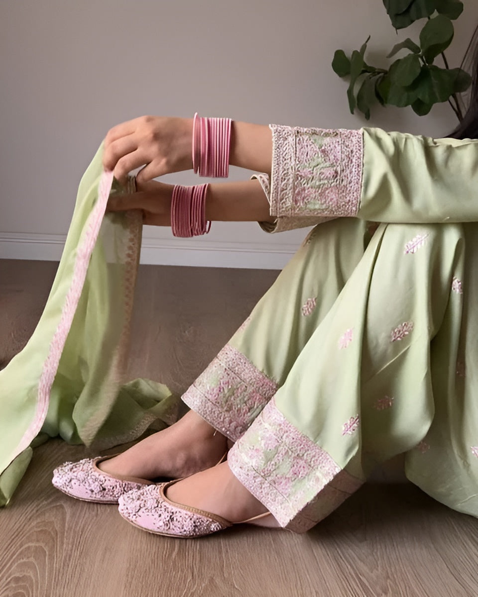 Person wearing a light green traditional outfit with pink embroidery, sitting on a wooden floor.