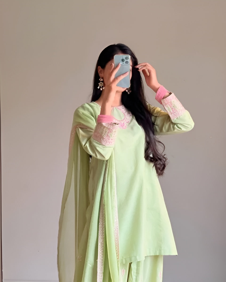 Person wearing a light green traditional outfit with pink embroidery, standing against a plain background.