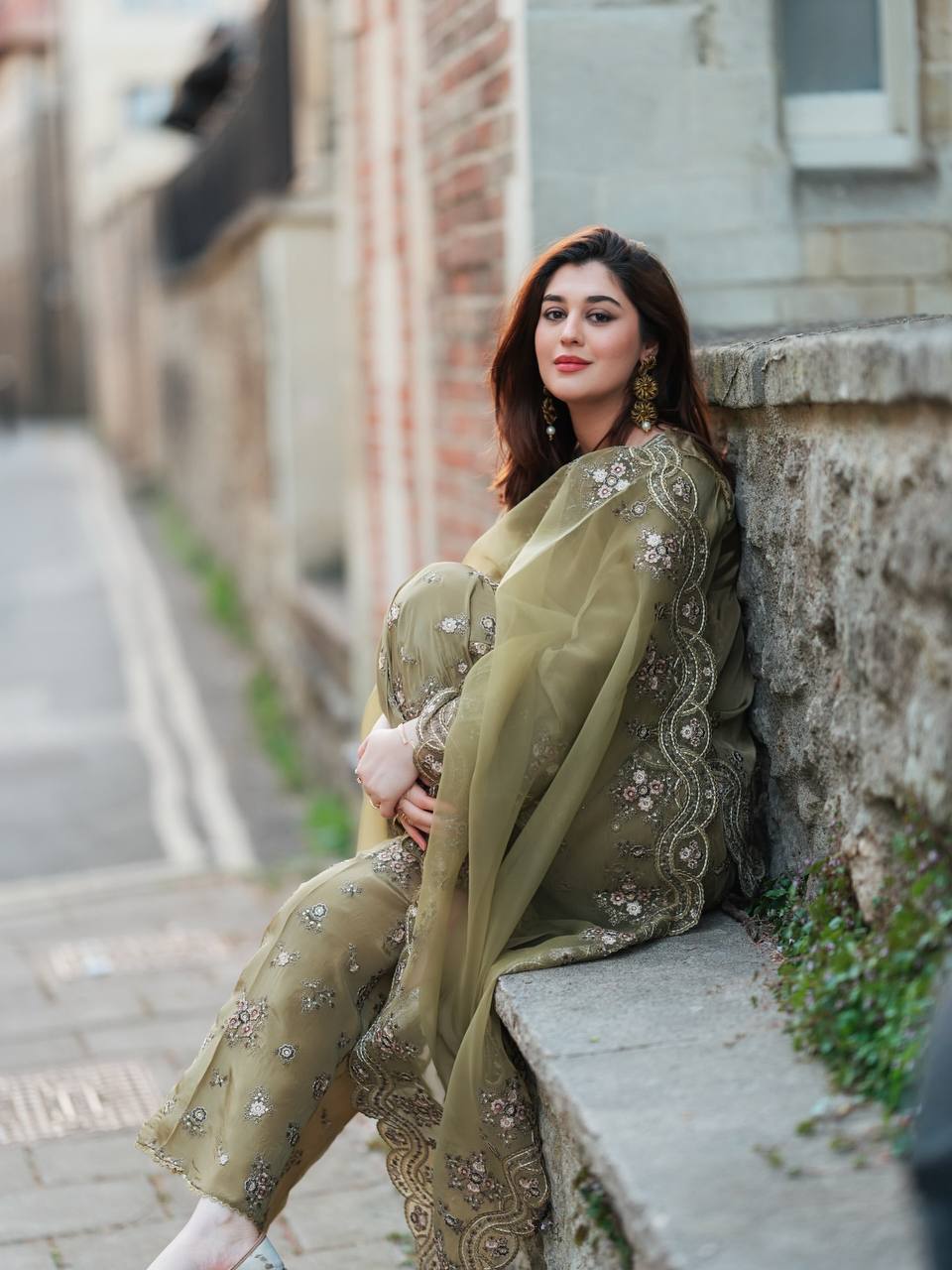Woman in a green traditional outfit sitting against a stone wall.