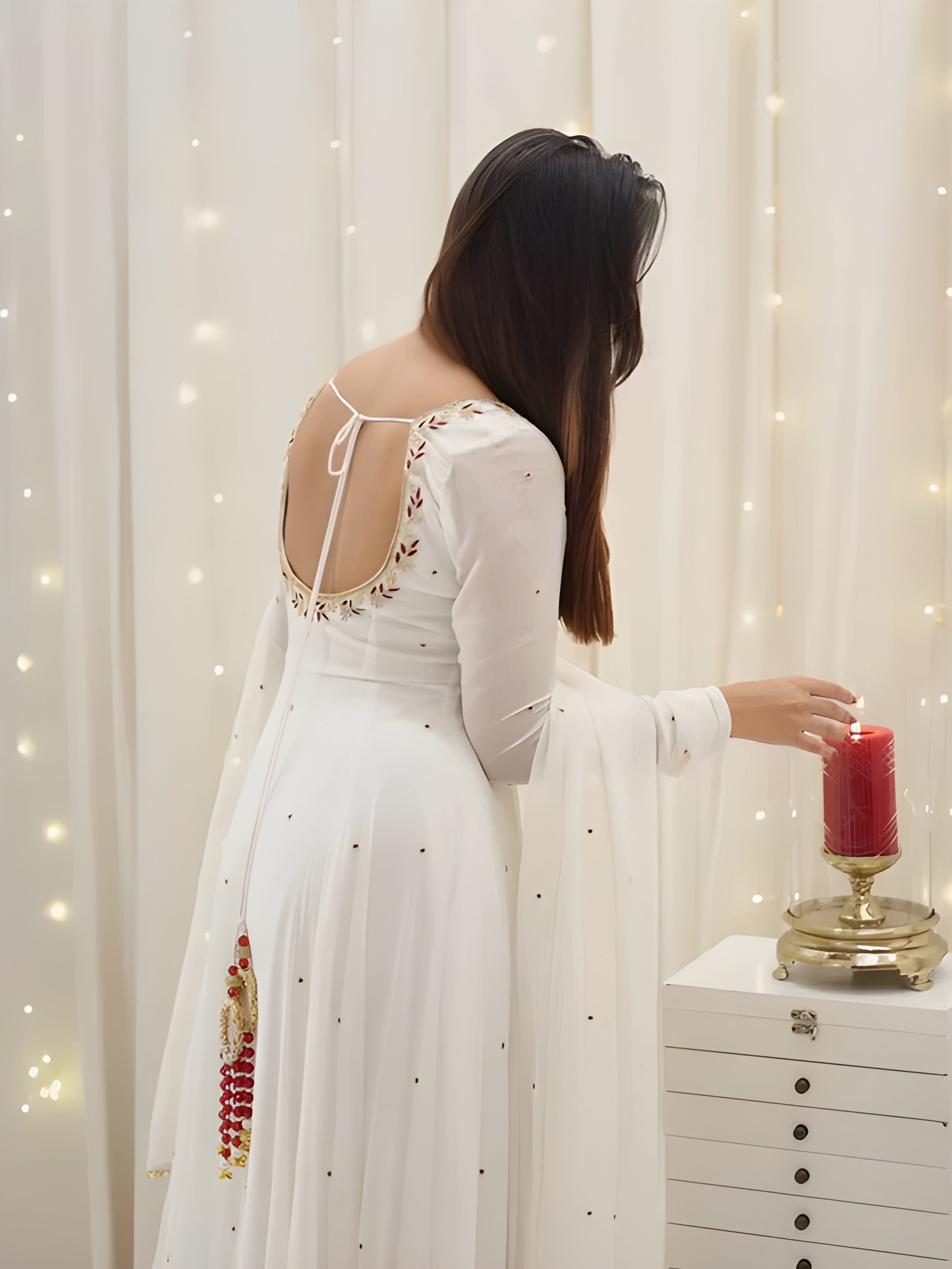Woman in a white dress with floral details standing in front of a white curtain with string lights.