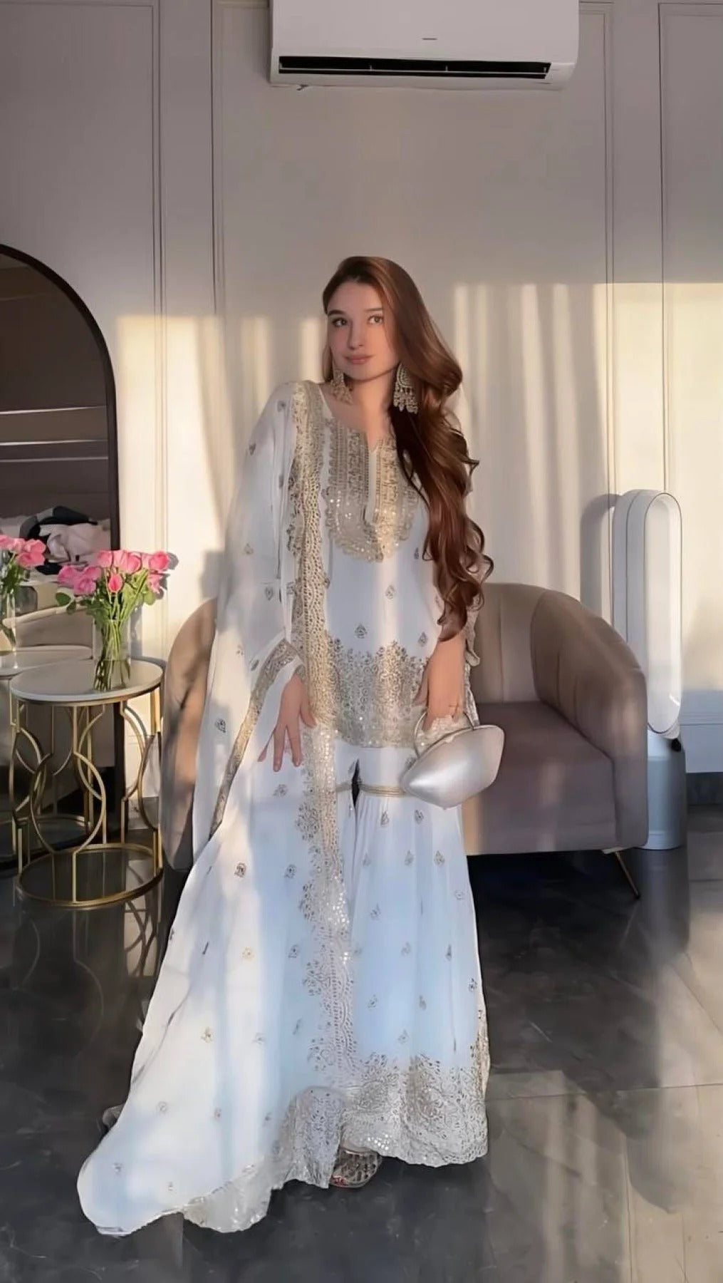 Woman in a light blue embroidered outfit standing in a modern interior setting.