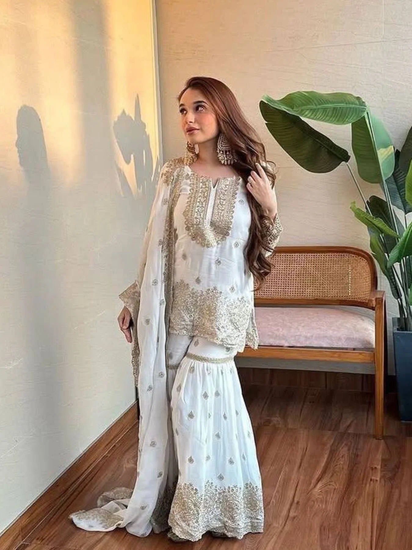 Woman in a white traditional outfit standing indoors with a plant and chair in the background
