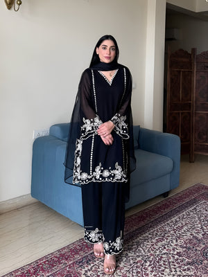 Woman in a black traditional outfit with white embroidery standing in a room with a blue couch and patterned rug.