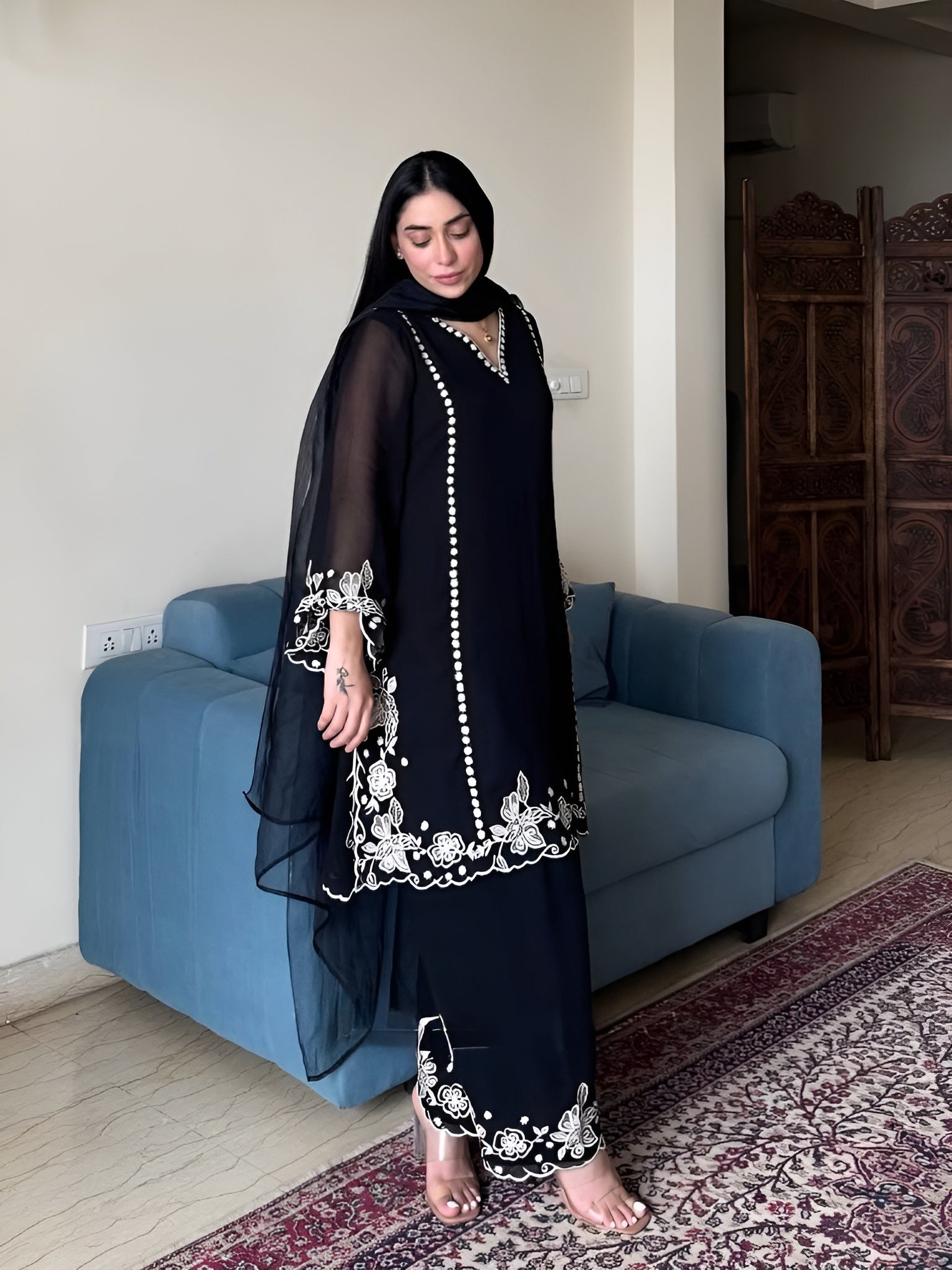 Woman in a black embroidered outfit standing in a room with a blue sofa and patterned rug.