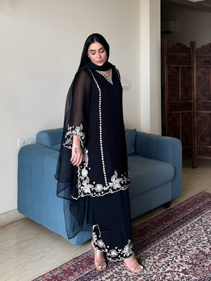 Woman in a black embroidered outfit standing in a room with a blue sofa and patterned rug.